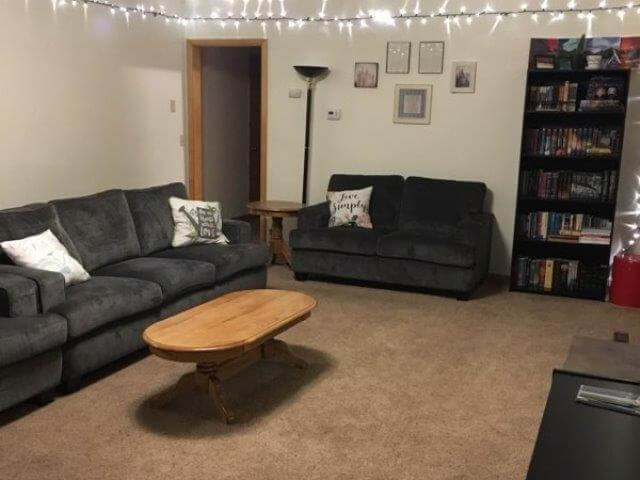 Apt #1 large living room showing grey sofa/loveseat, oak end tables, and residents’ decorative lights and bookshelf.