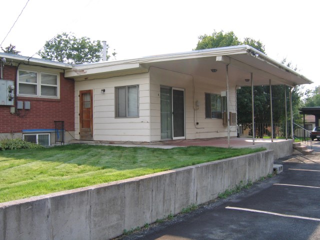 Exterior of house showing white siding, main door, sliding patio doors to large covered patio, green lawn, and parking.