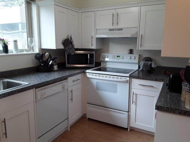 Kitchen with white cabinets, dark grey countertops, microwave, white stove, and large window. 